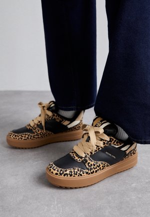 Person iført leopardmønstrede og sorte sneakers med tanfarvede såler og snørebånd, stående på en grå gulv med mørkeblå bukser.