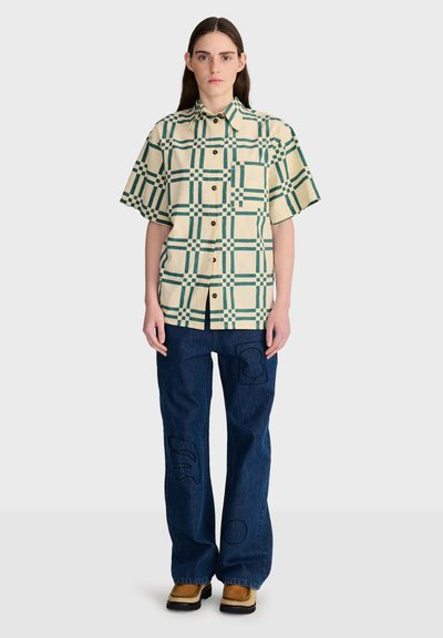 Femme debout portant une chemise beige à manches courtes avec un motif à carreaux verts, un jean bleu foncé avec des patchs et des chaussures marron.