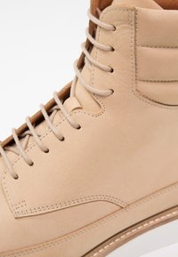 Beżowy skórzany but o gładkiej fakturze, usystematyzowanej konstrukcji, sześcioma oczkami na jasnoszare sznurowadła oraz kontrastującą białą podeszwą.