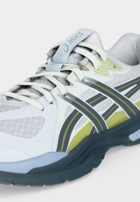 Λευκό και γκρι αθλητικό παπούτσι Asics με κίτρινες και μαύρες λεπτομέρειες, διχτυωτό επάνω μέρος, μαλακή σόλα και λευκά κορδόνια δεμένα μπροστά.