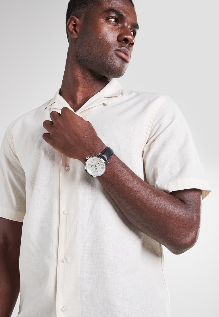 Montre pour homme avec un cadran rond argenté, un bracelet en cuir noir et des détails chronographes, portée par une personne dans une chemise à manches courtes beige clair.