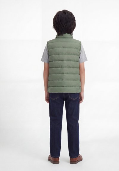 Παιδί που στέκεται με την πλάτη, φοράει πράσινο ενισχυμένο γιλέκο, γκρι κοντομάνικο μπλουζάκι, σκούρα τζιν και καφέ παπούτσια σε λευκό φόντο.