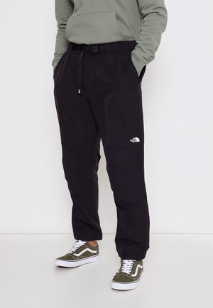 Person trägt schwarze The North Face Hose mit Seitentaschen und olivgrünen sowie weißen Sneakers, steht mit den Händen in den Taschen.