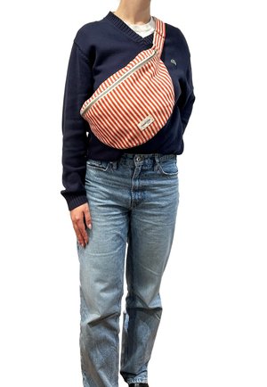 Personne portant un pull col en V bleu marine, un jean bleu clair et un sac bandoulière rayé rouge et blanc avec une fermeture éclair sur la poitrine.
