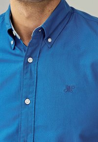 Camisa azul de botones con tejido texturizado, pequeños botones blancos y un logo bordado sutil en el pecho. Con cuello y ajuste clásico.