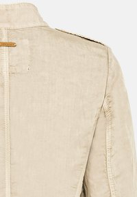 Dos de veste en tissu beige avec coutures surpiquées, épaulette et petite étiquette en cuir rectangulaire près de la couture supérieure gauche.