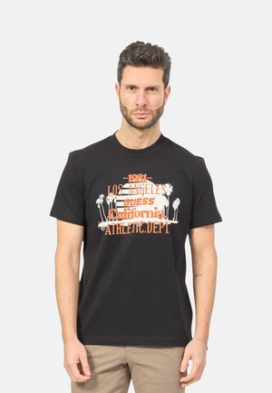 Man draagt een zwart T-shirt met oranje en witte tekst die "1981 Los Angeles Guess California Athletic Dept." leest en palmboom afbeeldingen.