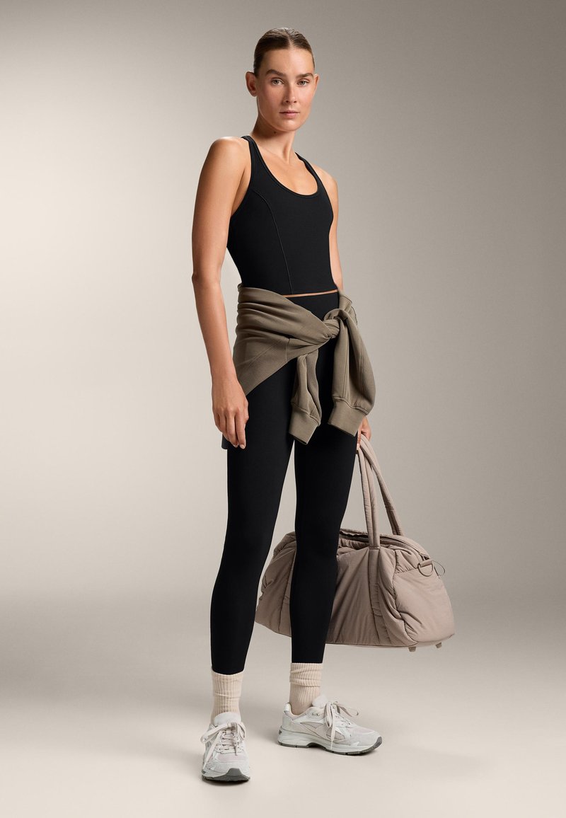 Musta urheilu toppi, jossa on tyylikäs muotoilu, yhdistettynä korkeavyötäröisiin mustiin leggareihin. Vaaleanharmaat tennarit ja beige kuntosalilaukku viimeistelevät ilmeen.