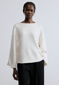 Krém színű, túlméretezett pulóver széles nyakkivágással, nyers szegéllyel és hosszú harang alakú ujjal, fekete nadrággal párosítva. Puha tapintású anyag.