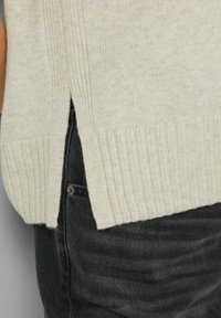 Beżowy sweter dzianinowy z prążkowanym dołem i bocznymi rozcięciami, wykonany z miękkiej tkaniny, w zestawieniu z ciemnymi dżinsami. Tekstura wydaje się przytulna i ciepła.