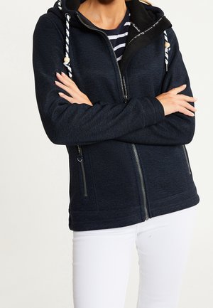 Personne portant un sweat à capuche bleu marine avec fermeture éclair et cordons blancs sur une chemise rayée et un pantalon blanc, les bras croisés sur la poitrine.