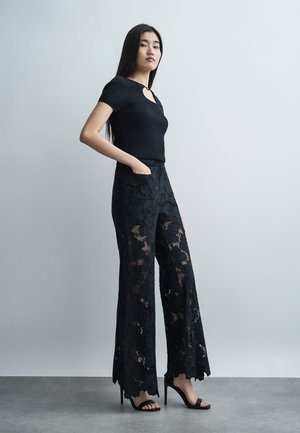 Kvinde står med hånden i lommen iført sort top med korte ærmer og vidbukser med blonde mønster samt høje hælede sandaler foran en lys væg.