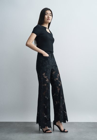 Femme debout, main dans la poche, portant un haut noir à manches courtes et un pantalon large à motifs en dentelle, avec des sandales à talons hauts, devant un mur clair.