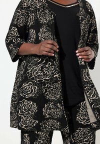 Mustapohjainen kukkakuosiset takki, joka on valmistettu tekstuurista kankaasta. Takissa on väljä malli, avomallinen etuosa ja kolme-neljäosa hihasia. Takki on puettu mustan paidan päälle.