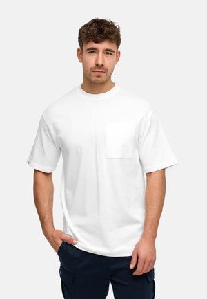 Jeune homme aux cheveux courts et bouclés portant un T-shirt blanc uni avec une poche et un pantalon foncé, debout avec une main dans la poche.