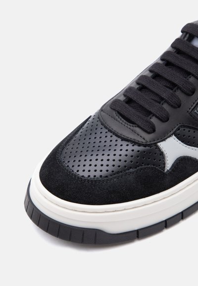 Baskets noir et blanc avec toe en cuir perforé, surpiqûres en suède, lacets noirs et semelle épaisse blanche sur fond uni.