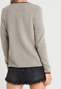 Haut à manches longues beige avec un motif hexagonal noir, doté d'un col rond et d'une texture douce, associé à un short en denim noir.