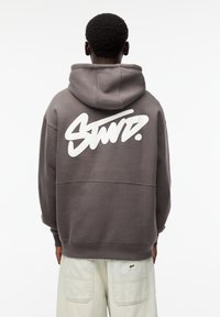Sudadera gris con un logotipo gráfico blanco centrado en la parte posterior. Presenta una capucha amplia y puños acanalados. Tejido suave y texturizado.