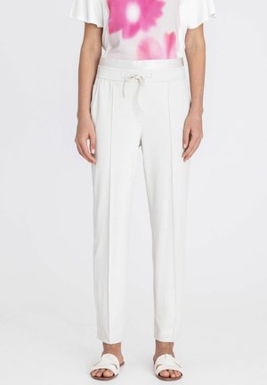 Vrouw draagt witte broek met hoge taille en voorplooien, met een koord in de taille, gecombineerd met witte slippers en een roze-wit topje.