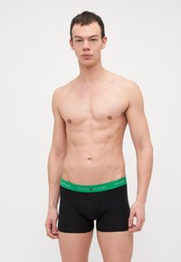 Schwarze Boxershorts mit grünem Taillenbund, auf dem "TOMMY HILFIGER" steht; glattes Material und körpernahe Passform für Komfort.