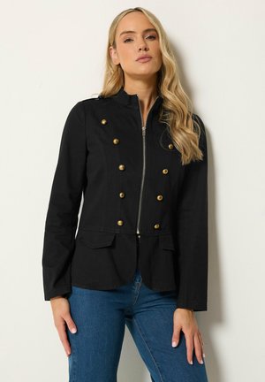 Frau mit schwarzer Militärjacke mit goldenen Knöpfen und blauer Jeans steht vor einem schlichten weißen Hintergrund.