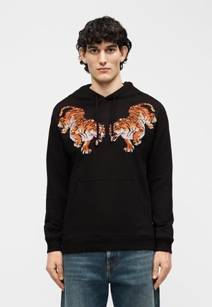 Jonge man met donker krullend haar die een zwarte hoodie draagt met daarop twee oranje-witte tijgers die elkaar in de borststreek spiegelen.
