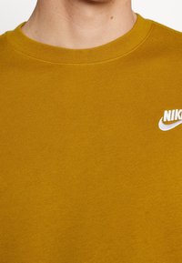 Gul bomullströja med rundad halsringning. Har en vit Nike-logga på vänster bröstområde. Slät textur, avslappnad design.