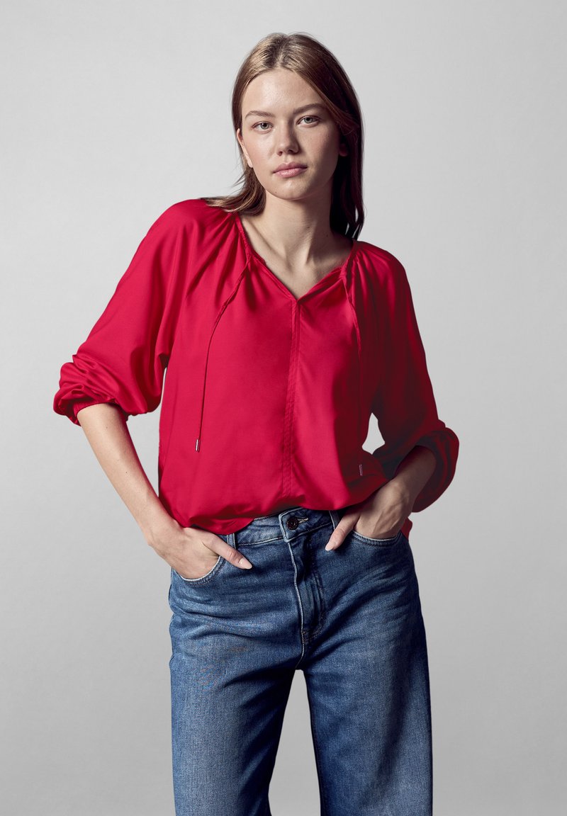 Blusa rossa a maniche lunghe con scollatura a V e dettaglio con cordino. Abbinata a jeans in denim blu. Tessuto liscio, vestibilità rilassata.