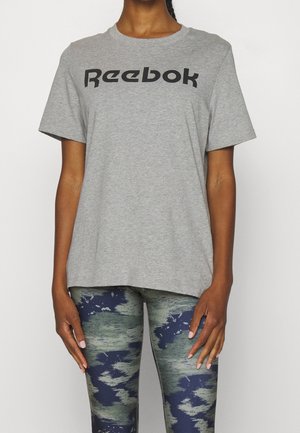 Persona con una camiseta gris de Reebok y leggings con patrón de camuflaje, de pie frente a un fondo blanco liso.