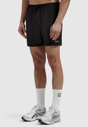 Man draagt zwarte sportshorts, witte sokken met zwarte tekst en zilveren hardloopschoenen, staand tegen een effen achtergrond.