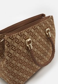 Brązowa torba tote z wzorem, z złotym okuciem, wytwornymi brązowymi uchwytami i wyraźną konstrukcją. Z przodu znajduje się logo w złotej literze.