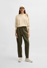 Beige sweatshirt met strikdetail, olijfgroene elastische broek en beige sneakers. Zachte stof, losse pasvorm en zichtbare stikselaccenten.