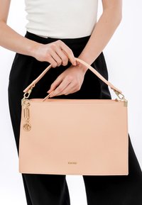 Naine hoiab heleroosat nahast käekotti, millel on kuldsed detailid ja logo, kannab valget toppi ja musti pükse valgel taustal.