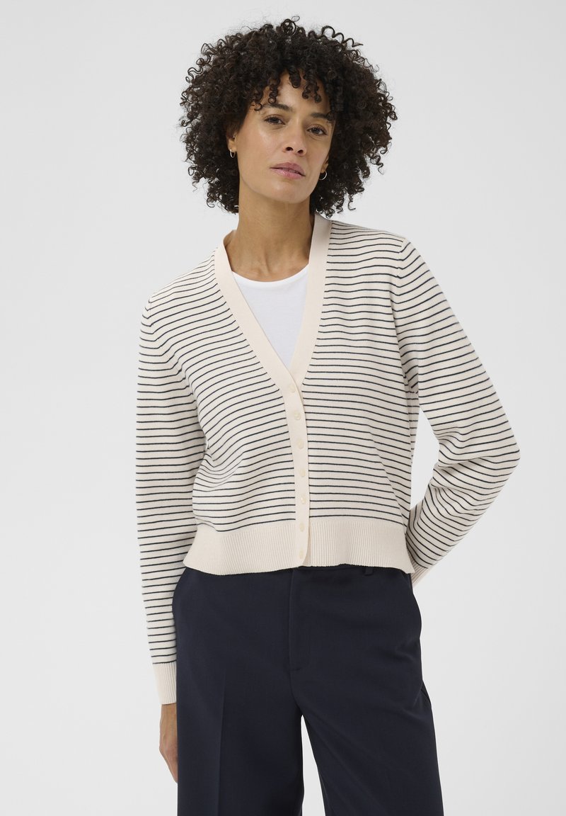 Femme aux cheveux bouclés portant un cardigan crème boutonné avec de fines rayures horizontales noires, par-dessus un haut blanc et un pantalon bleu marine foncé.