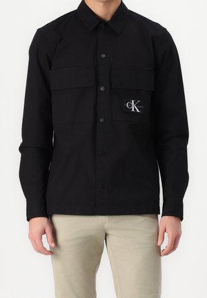 Mężczyzna w czarnej koszuli na guziki z dwoma kieszeniami na piersi, na jednej widnieje logo Calvin Klein Jeans, w połączeniu z beżowymi spodniami.