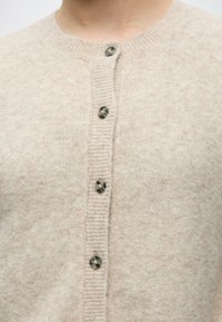 Nahaufnahme eines beigen Strickcardigans mit geripptem Saum und fünf marmorierten Knöpfen vorne, getragen von einer Person.
