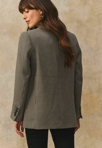 Blazer med sort og hvidt houndstooth-mønster, lange ærmer, slim fit og knaplukning ved ærmerne, vist fra bagsiden.