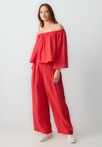Haut rouge à épaules dénudées associé à un pantalon large assorti, le tout en tissu léger avec une finition texturée, complété par des baskets blanches.