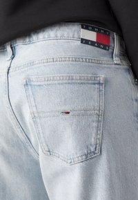 Světle modré džíny z denimu s vybledlým povrchem. Obsahuje zadní kapsu s malou červenou, bílou a modrou logovou nášivkou a žlutými švy.