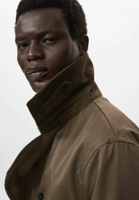 Bruine trenchcoat met hoge kraag, voorzien van grote knopen en een soepele textuur, model draagt deze en staat lichtjes naar links gericht tegen een witte achtergrond.