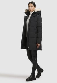 Sort pufferjakke med hætte foret med pels, lynlås og sidelommer. Båret over en hvid top og parret med sorte leggings og ankelstøvler.
