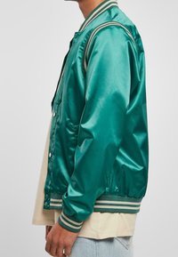 Türkisfarbige Satin-Bomberjacke mit geripptem grün-weiß gestreiftem Kragen und Ärmelbündchen. Mit Druckknöpfen, Seitentaschen und glatter Textur.