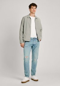 Chaqueta gris claro con cuello, llevada sobre una camiseta blanca. Jeans rasgados de color azul claro y zapatillas blancas con suelas marrones.