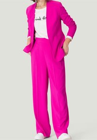 Veste blazer ajustée rose vif et pantalon à jambes larges, portés par-dessus un t-shirt blanc avec du texte noir. Tissu lisse avec un design structuré.