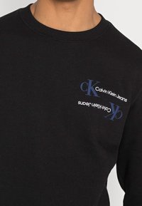 Černý svetr vyrobený z bavlny, s kulatým výstřihem. Na levé části hrudi je modrý potisk: "Calvin Klein Jeans" a "CK".