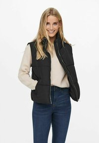 Gilet noir matelassé avec fermeture éclair sur le devant et poches latérales, porté par-dessus un pull beige, assorti à un jean bleu taille haute.