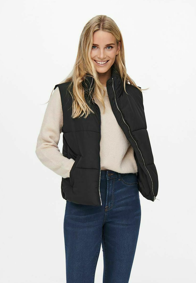 Gilet noir matelassé avec fermeture éclair sur le devant et poches latérales, porté par-dessus un pull beige, assorti à un jean bleu taille haute.