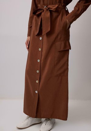 Jupe marron à boutons sur le devant avec ceinture nouée à la taille et poche à rabat latérale, portée avec des chaussures blanches à lacets.