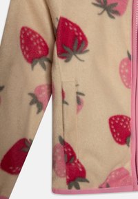 Jachetă din fleece cu un model de căpșuni roz pe un fundal bej, cu o buzunăraș pe laterale și bordură roz de-a lungul fermoarului. Textură moale.