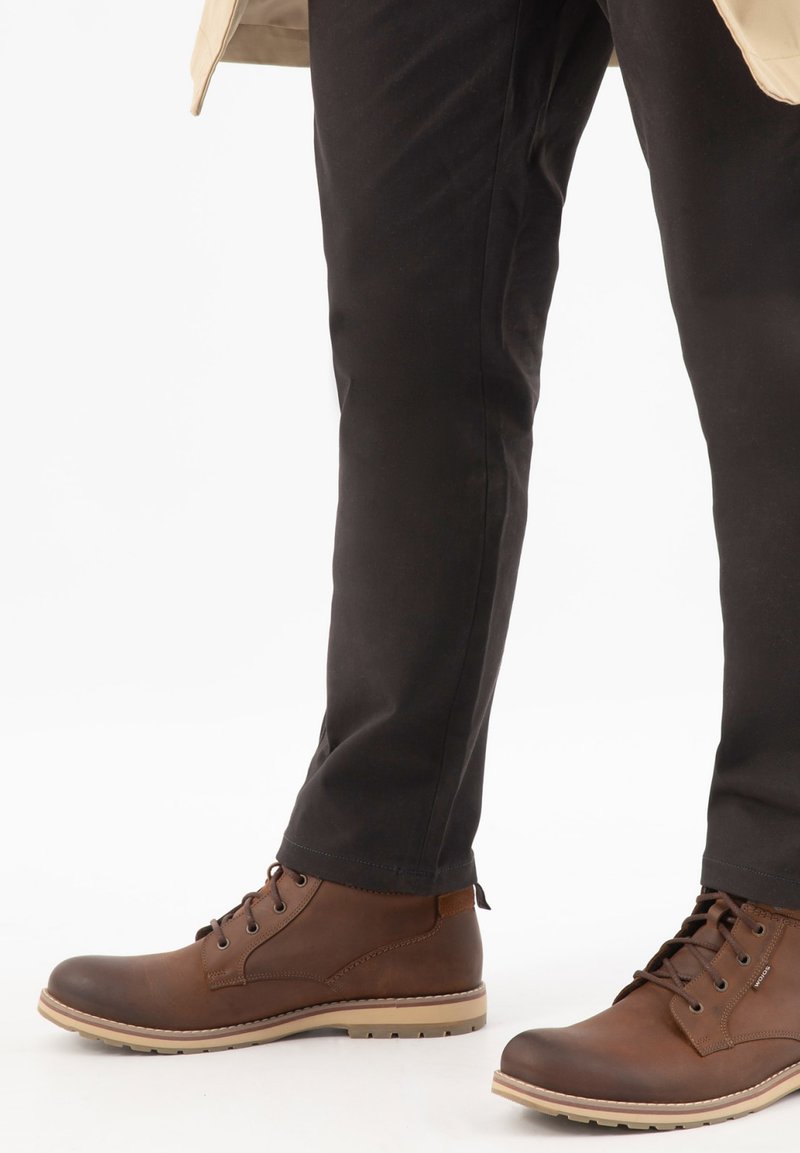 Bottes en cuir marron à lacets portées avec un pantalon noir et un ourlet de manteau beige visible en haut.
